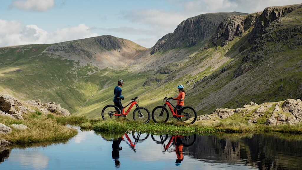 Zwei Mountainbiker mit roten Rädern in alpiner Landschaft, reflektiert in einem klaren Bergsee, diskutieren über Fahrräder.