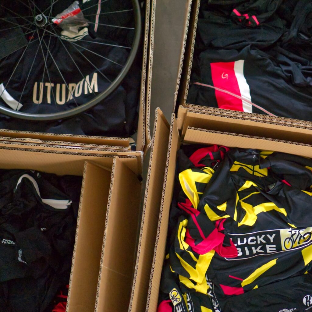 Kartonboxen mit bunten Radtrikots und einem Fahrradreifen, bereit für den Versand in Düsseldorf.