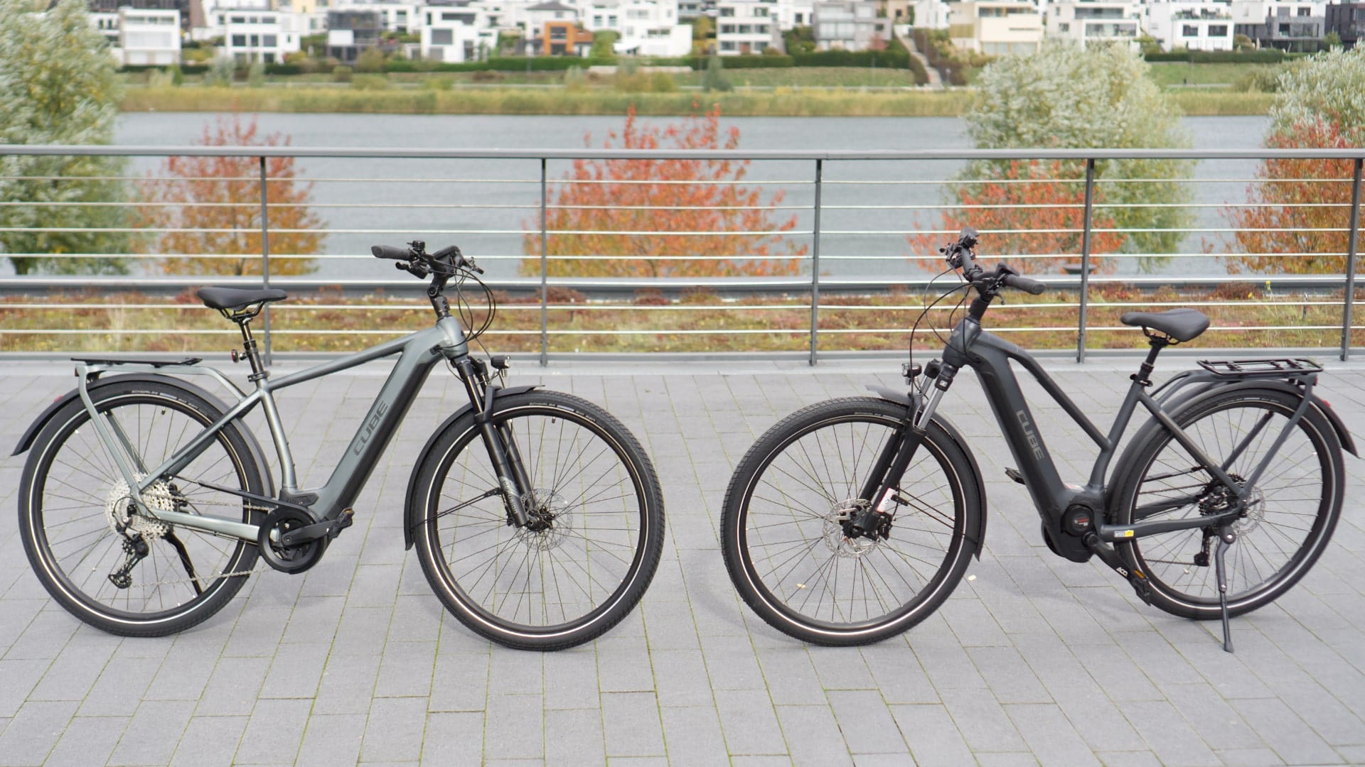 Zwei Cube Kathmandu Hybrid 625 E-Bikes im Vergleich, nebeneinander auf einer Promenade vor herbstlicher Kulisse.