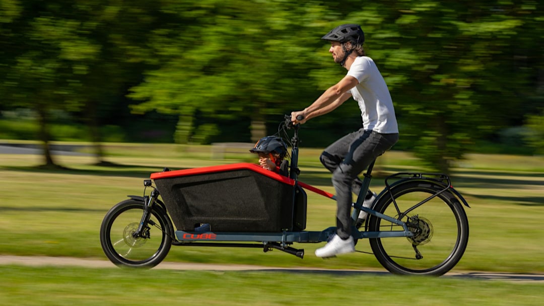 Mann fährt mit einem Cube E-Lastenrad im Park; Kind sitzt sicher im vorderen Transportbereich.