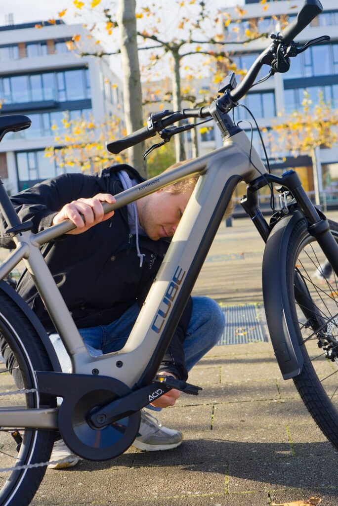 Mann überprüft Akku eines Cube Kathmandu Hybrid 750 E-Bikes im Freien, moderne Architektur im Hintergrund.