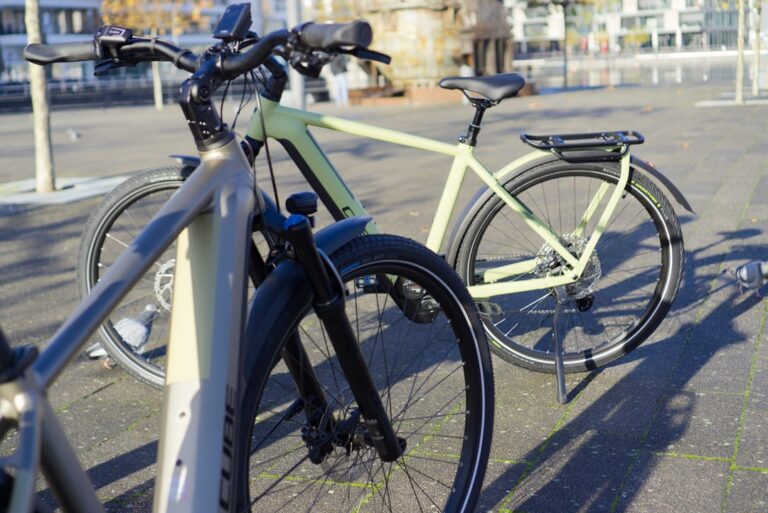 Zwei Cube Kathmandu Hybrid 750 E-Bikes im Vergleich auf städtischem Platz bei Sonnenschein.