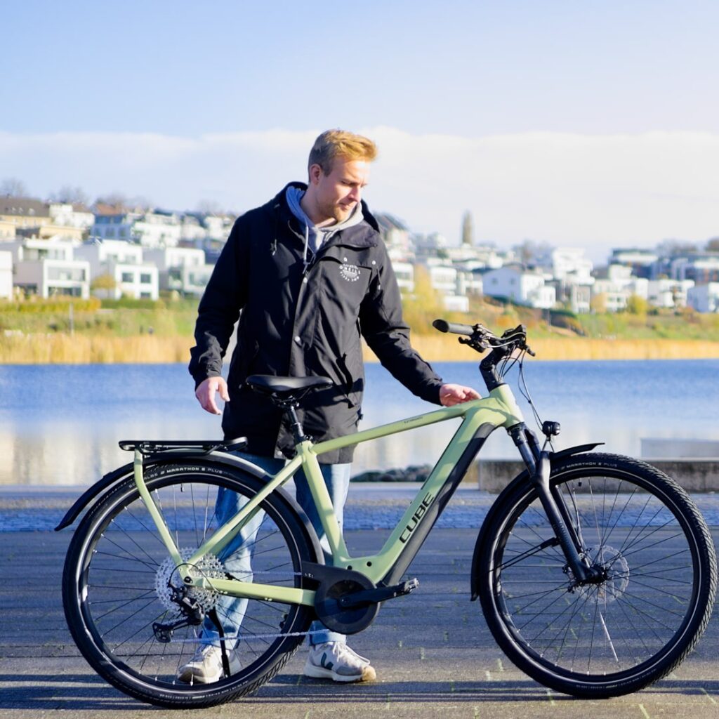 Mann neben Cube Kathmandu Hybrid Fahrrad am Seeufer, moderne Architektur im Hintergrund.