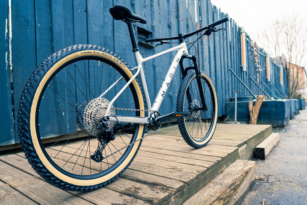 Marin Team Marin 1 2023 Mountainbike auf Holzplattform vor blauer Wand, zeigt robuste Bauweise und Offroad-Bereifung.