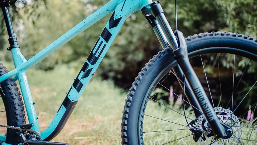 Trek Roscoe 7 2023 Rahmen in Nahaufnahme, türkisfarben, mit robusten Reifen und RockShox-Gabel im Grünen.