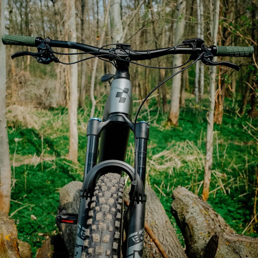 Cube Stereo Hybrid 140 HPC TM 750 2023 im Wald, Vorderansicht, Fokus auf Lenker und Vorderrad, robustes E-Mountainbike.