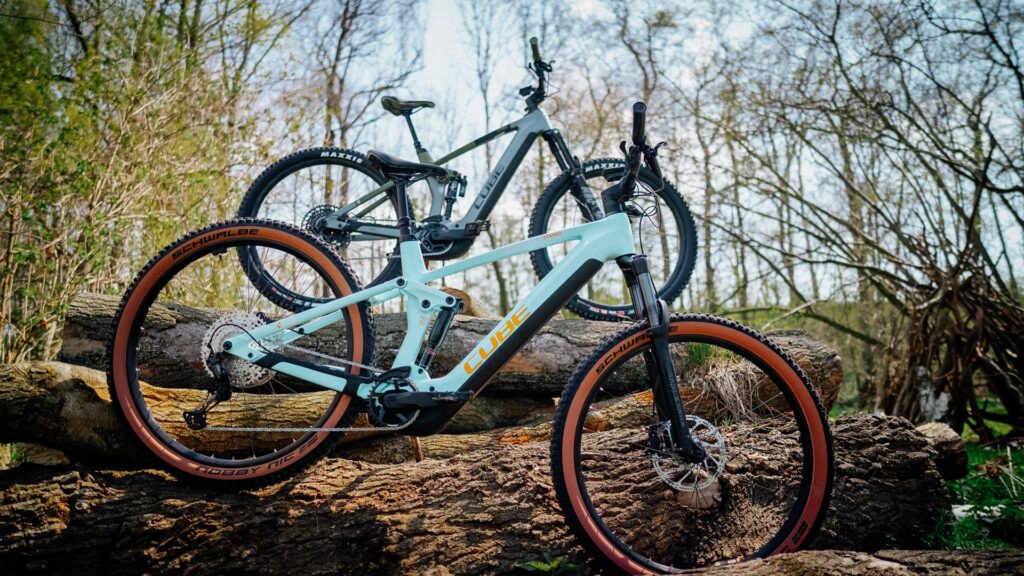 Zwei Cube Stereo Hybrid 140 HPC E-Mountainbikes im Wald auf Baumstämmen präsentiert.