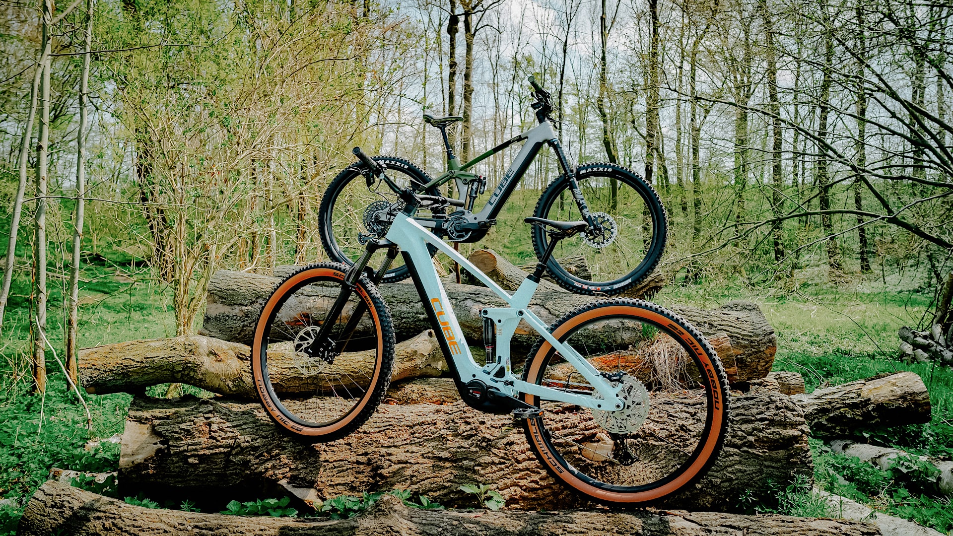 Zwei Cube Stereo Hybrid 140 HPC 750 E-Mountainbikes im Wald auf Baumstämmen.
