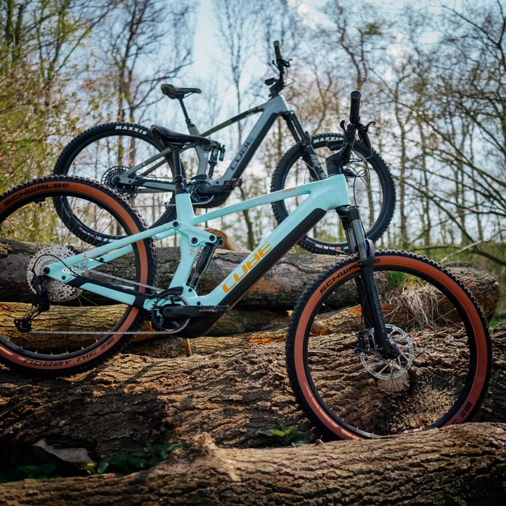 Zwei Cube Stereo Hybrid 140 HPC E-Mountainbikes im Wald auf Baumstämmen, Modellvergleich 2023.