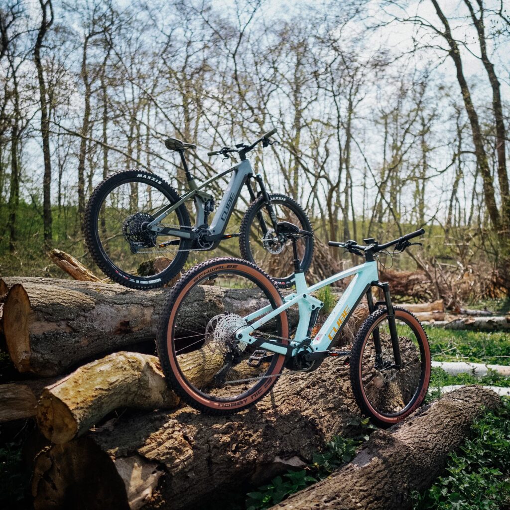 Zwei Cube Stereo Hybrid 140 HPC 750 E-Mountainbikes auf Baumstämmen im Wald, von hinten betrachtet.