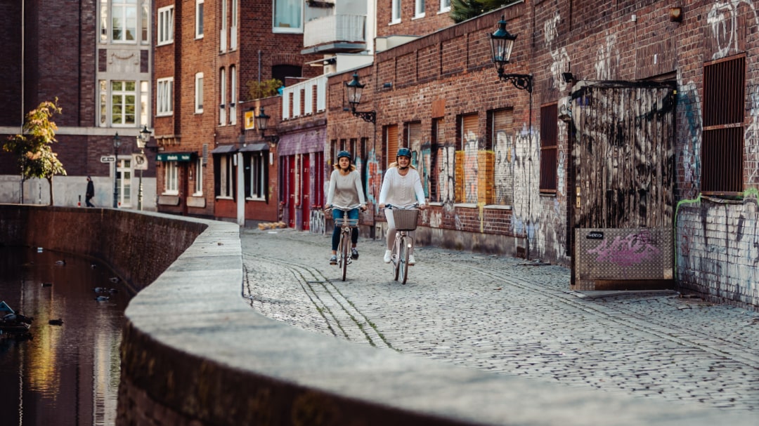 Zwei Personen radeln auf Citybikes entlang eines gepflasterten Wegs in urbaner Umgebung mit Graffiti-Wänden.