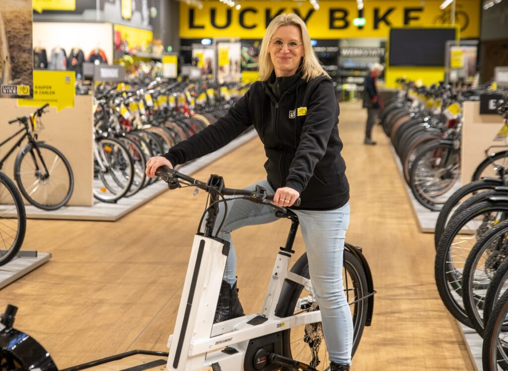 Mitarbeiterin auf E-Lastenrad in Fahrradgeschäft, umgeben von Fahrrädern, lächelnd in die Kamera.