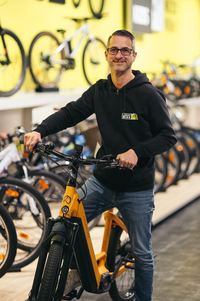 Mitarbeiter mit E-Citybike im Fahrradgeschäft, umgeben von weiteren Fahrrädern, lächelnd in die Kamera.