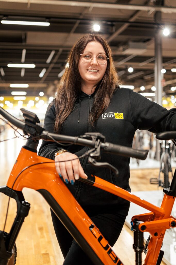 Mitarbeiterin präsentiert orangefarbenes E-Fully in einem Fahrradgeschäft, passend zum E-Bike-Guide-Thema.