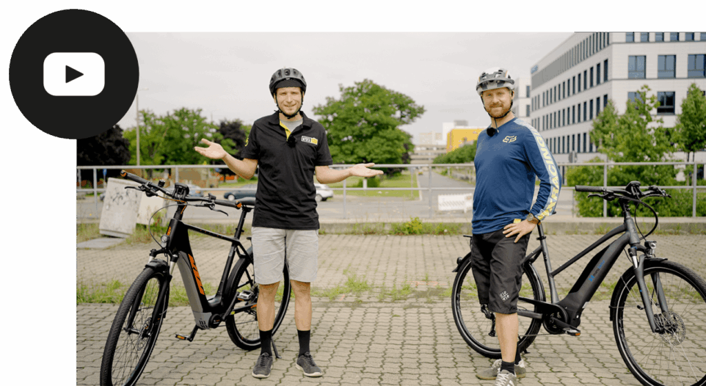 Zwei Männer mit Helmen präsentieren E-Bikes im Freien, umgeben von modernen Gebäuden.