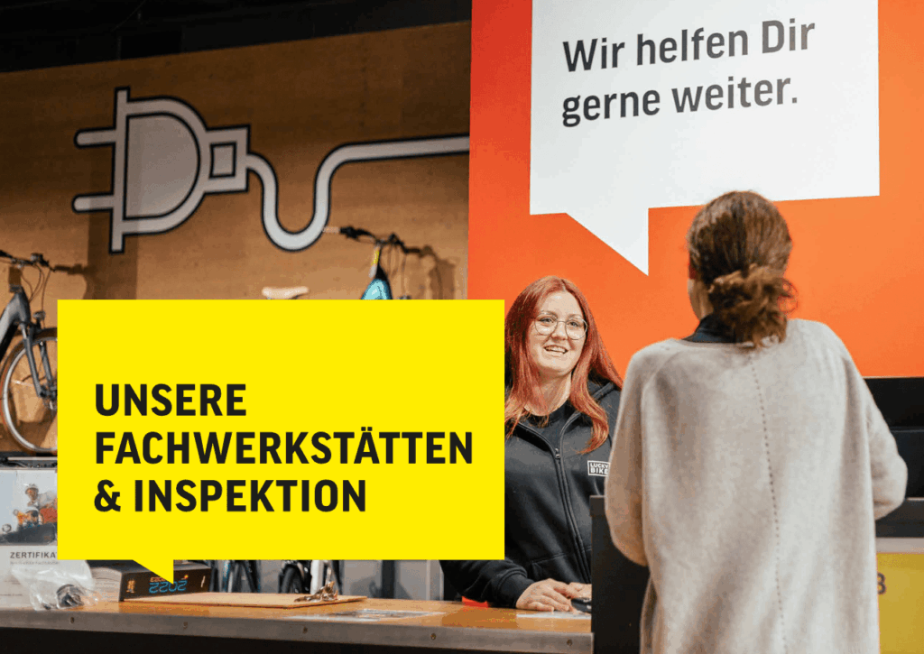 Fachwerkstatt-Mitarbeiterin berät Kundin zur E-Bike-Inspektion, gelbes Schild mit Text im Vordergrund.