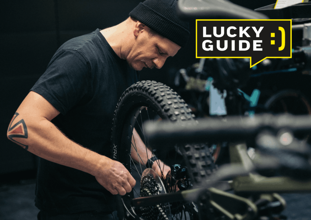 Mechaniker repariert E-Bike in Werkstatt, Fokus auf Hinterrad und technische Details, mit Lucky Guide-Logo.