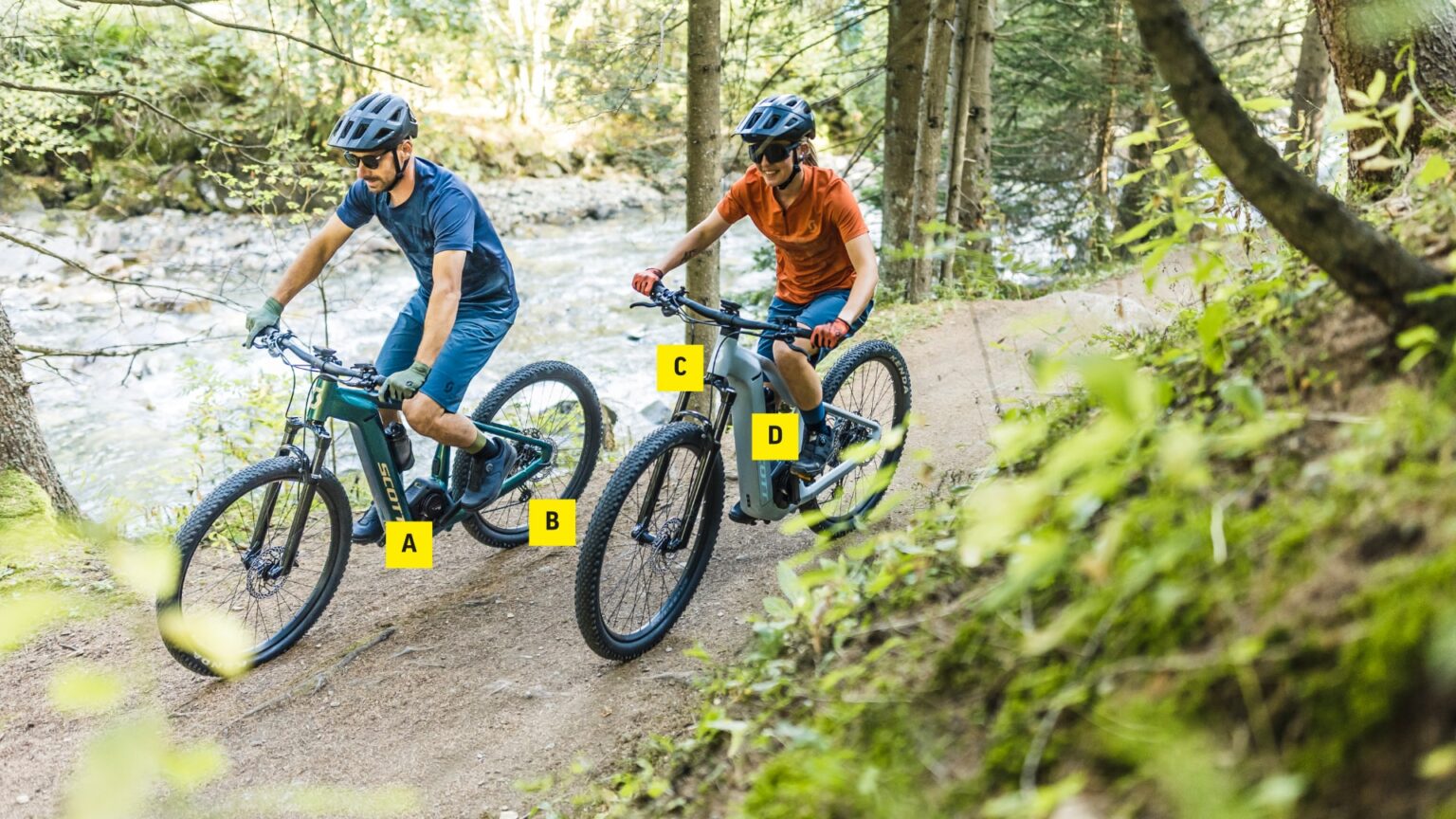 Zwei Radfahrer fahren auf einem E-Mountainbike-Hardtail durch einen bewaldeten Pfad am Fluss.