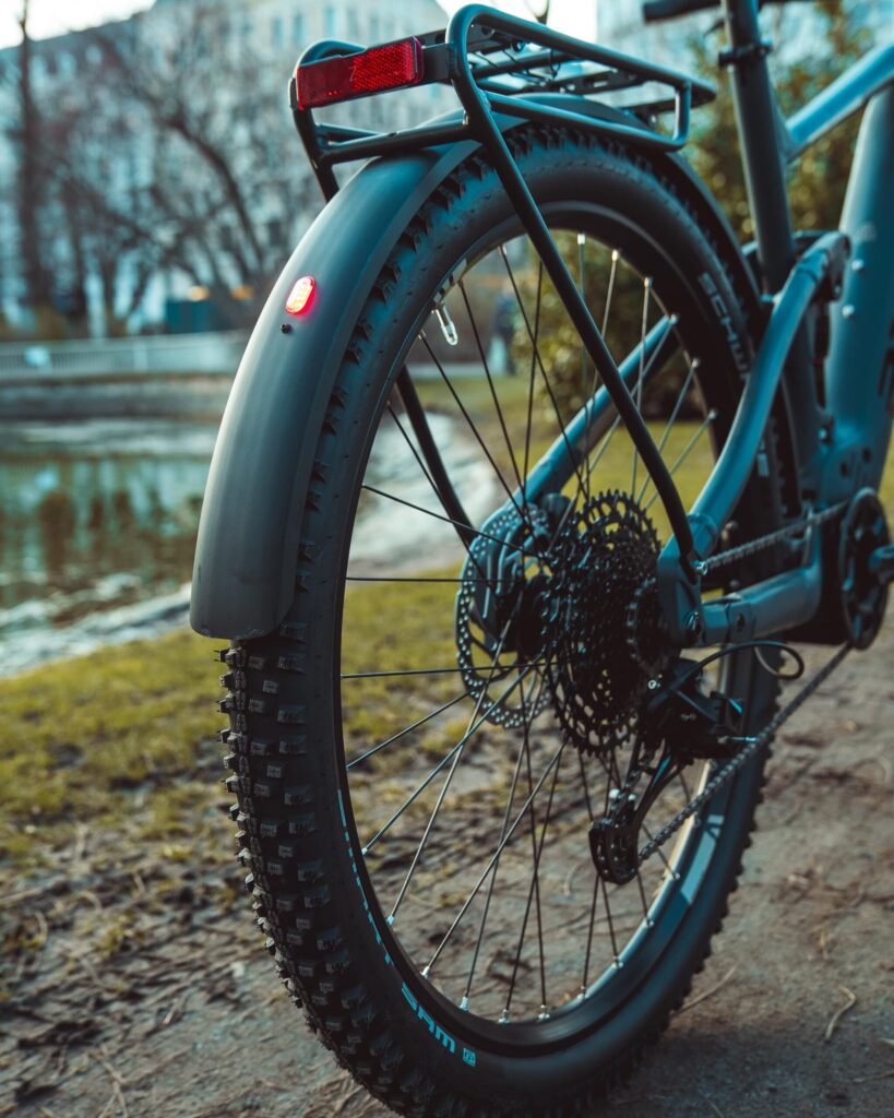 Hinterrad des Axess Rogue FS Allroad 2024 mit Schutzblech und Gepäckträger, parkend auf einem Weg am Wasser.