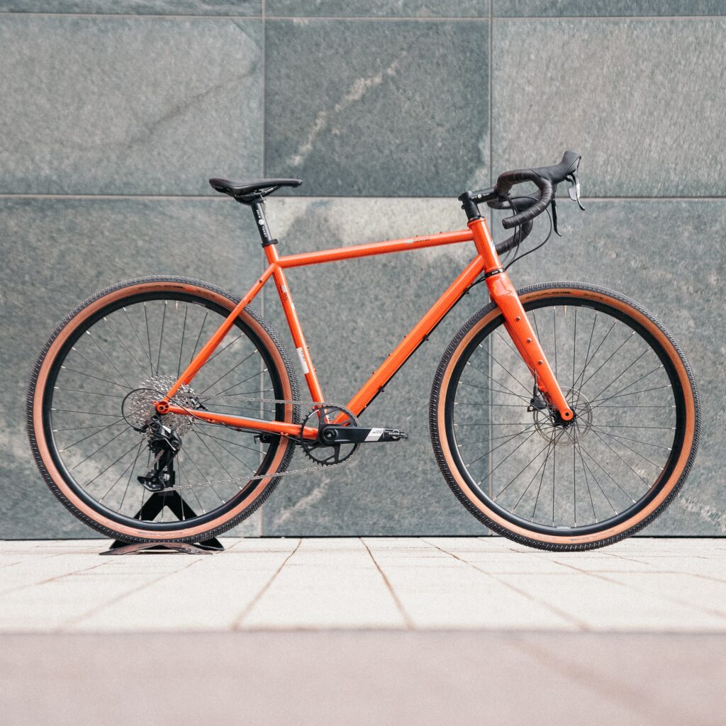Orangefarbenes VSF GX-500 Fahrrad vor grauer Wand, seitliche Ansicht, betont modernes Design und Funktionalität.