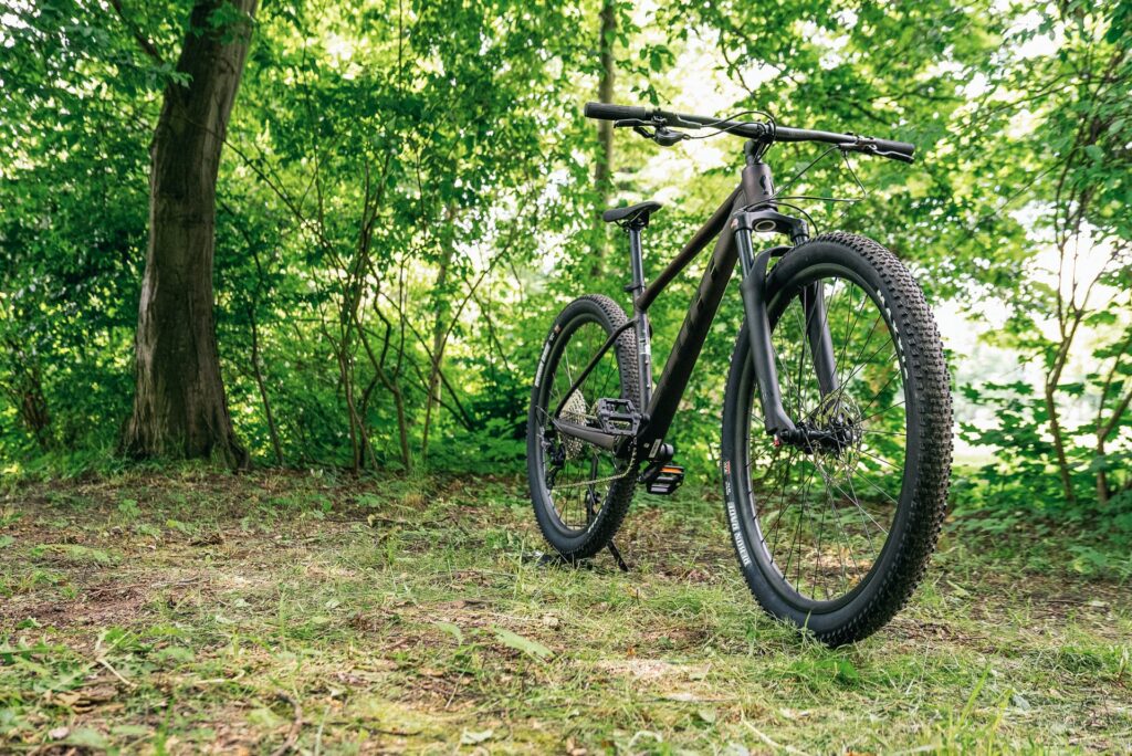 Scott Scale 980 2024 Mountainbike im Wald, seitliche Frontansicht, betont robuste Reifen und schlankes Design.