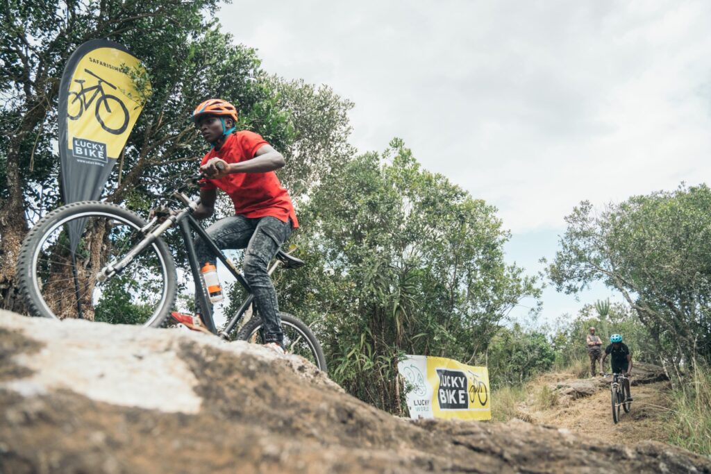 Mountainbiker auf Waldpfad bei Safari Simbaz Event, mit Lucky Bike Banner im Hintergrund.