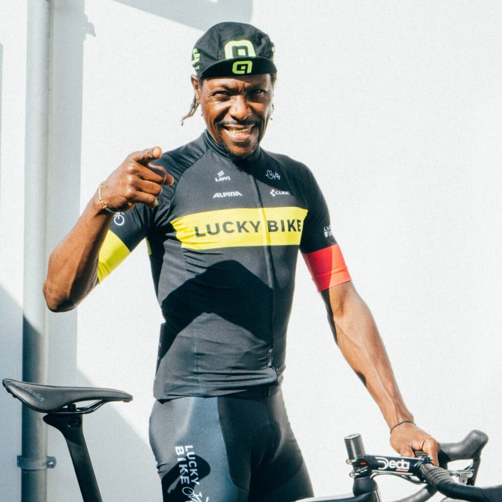David Kinjah in Lucky-Bike-Kleidung lächelt und zeigt auf Kamera, neben Fahrrad stehend, symbolisiert Abenteuer und Entschlossenheit.