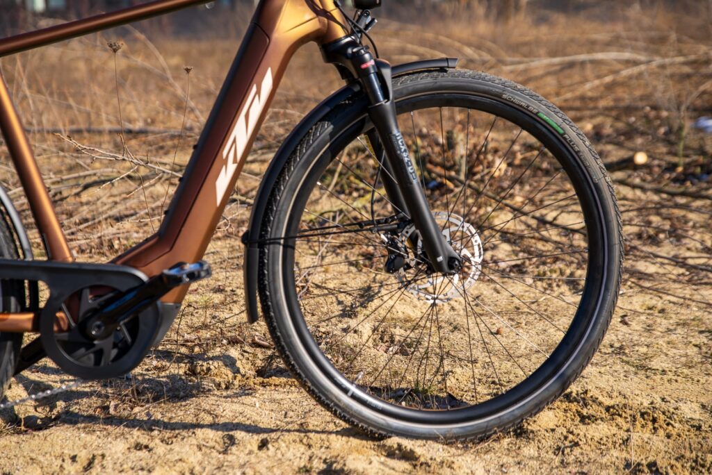 KTM MACINA Style Fahrrad, Nahaufnahme des Vorderrads mit Scheibenbremse auf sandigem Untergrund.