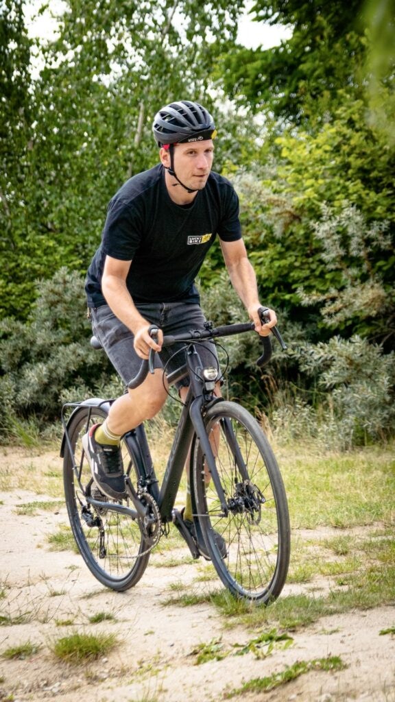 Mann fährt Gravelbike Axess Creed auf sandigem Weg, umgeben von grüner Natur, im Rahmen eines Vergleichstests.