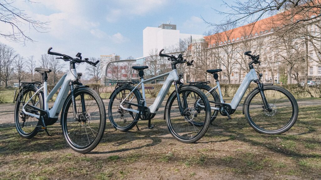 Drei KTM Macina Style X Pro 2025 E-Bikes im Park vor städtischer Kulisse, bereit für den Test.
