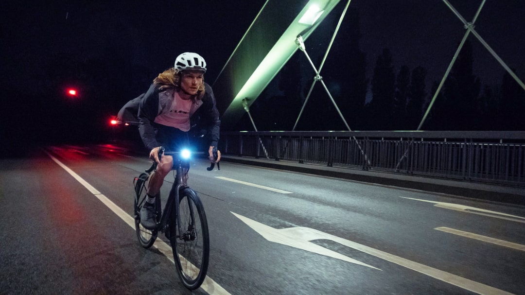Rennradfahrer bei Nacht auf beleuchteter Brücke, trägt Helm und reflektierende Kleidung, Cube Nuroad FE.