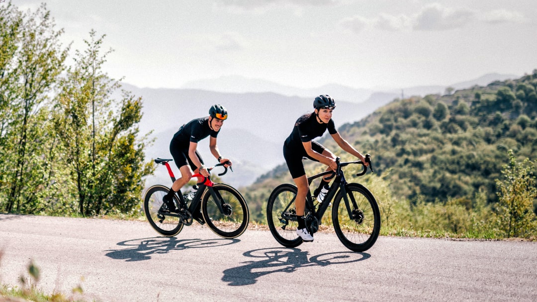 Zwei Radfahrer beim Bergfahren auf Rennrädern in sonniger Landschaft.