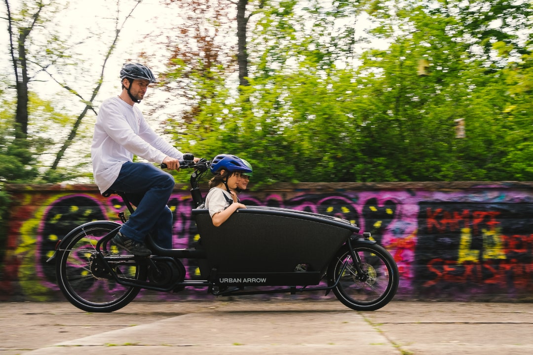 E-Lastenrad Urban Arrow Family mit Erwachsenem und Kind in urbanem Umfeld. Ideal für nachhaltige Mobilität.
