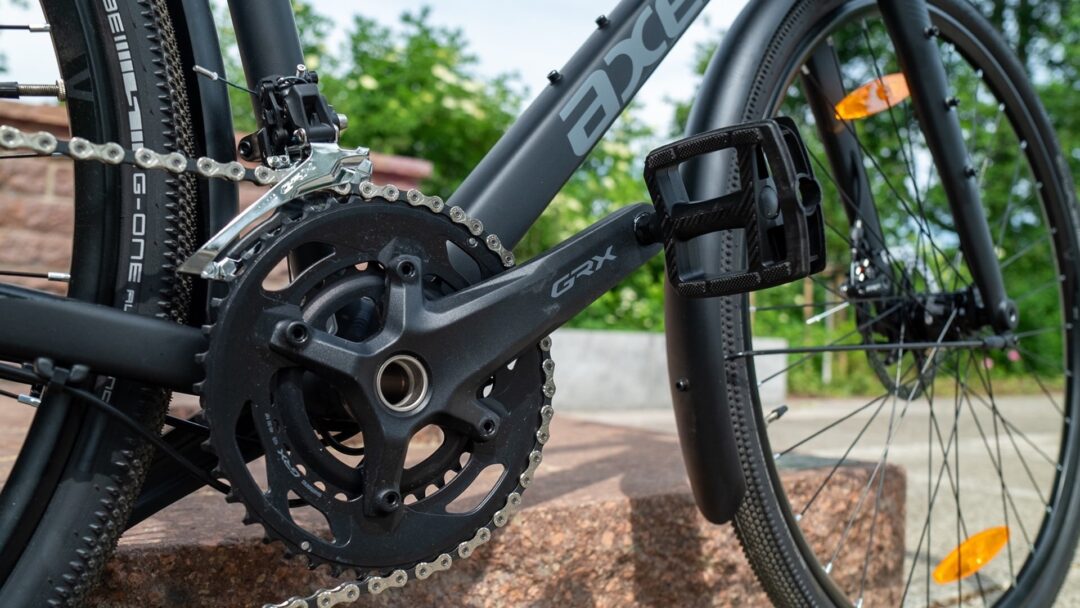 Detailaufnahme eines Axess-Gravelbikes: Shimano GRX-Kurbel mit 2-fach-Kettenblatt, Umwerfer, Plattformpedal, Schutzblech und Schwalbe G-One Allround-Reifen