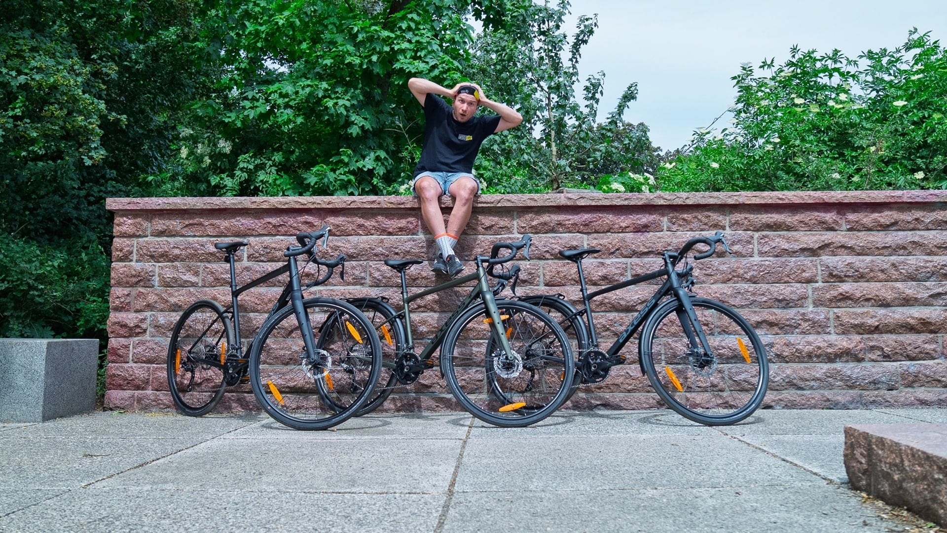 Vier Axess-Gravelbikes mit Dropbar und Scheibenbremsen vor einer Mauer; darüber sitzt eine Person mit Händen am Kopf auf der Mauer