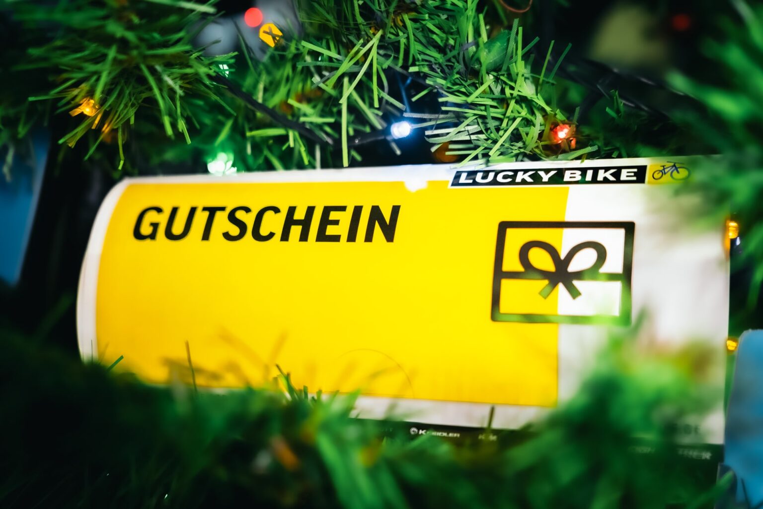 Lucky Bike Geschenkgutschein mit gelbem Hintergrund zwischen grünem Weihnachtsbaum und bunten LED-Lichtern