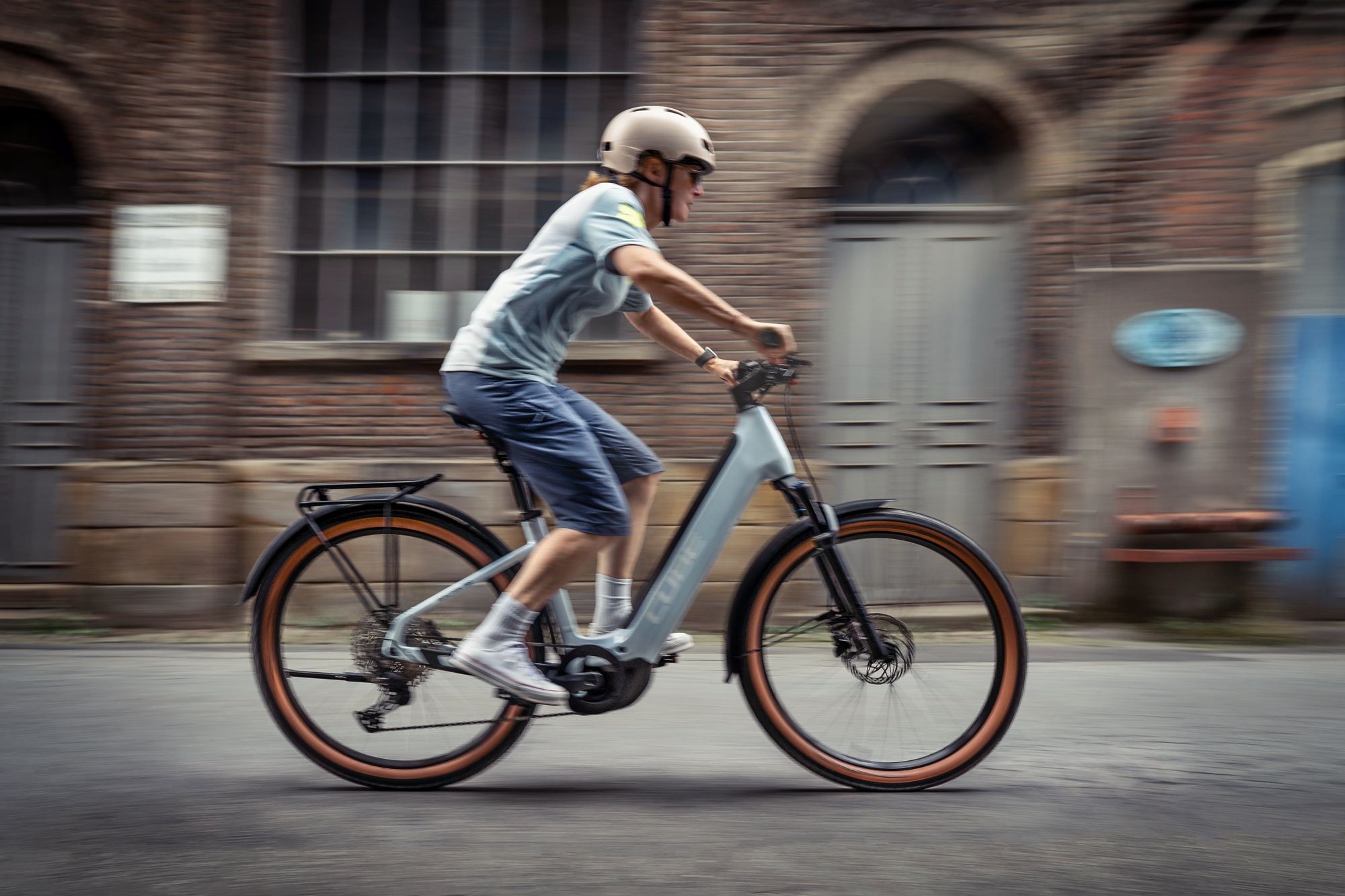 Frau auf einem E-Bike fährt von links nach rechts durch das Bild, im Hintergrund ist eine Hauswand mit Fenster und Tür zu sehen.
